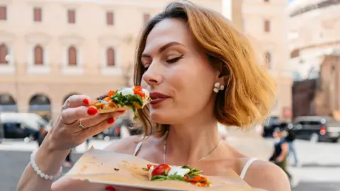Getty Images A woman eats a slice of pizza on the street in Rome. She has short hair.
