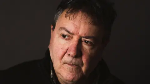 A headshot of Oscar McLennan, a man with darlk hair and wearing a dark top. He is sitting down, with an acoustic guitar 