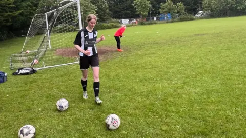 A football player from Odd Down Women's AFC wearing a black and white striped top and black shorts goes towards a football near the goal on a pitch. 