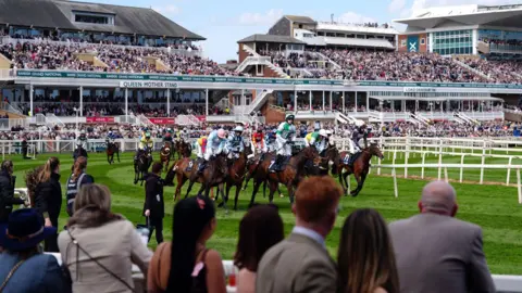 PA Media A group of horses and jockeys finish a race in their multicoloured silks in front of crowded stands at the Grand National meeting
