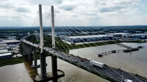 The large QEII Bridge that crosses the River Thames. It has two large beams with lots of long support wires holding it up. Cars and lorries are crossing in this picture.
