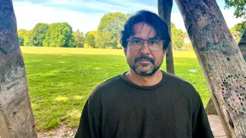 Hugo in a dark olive T-shirt sitting on a bench that is part of a wooden structure in a park in Exeter. He has dark hair and a beard that is flecked with some grey and he is wearing glasses. Hugo has a serious expression on his face.