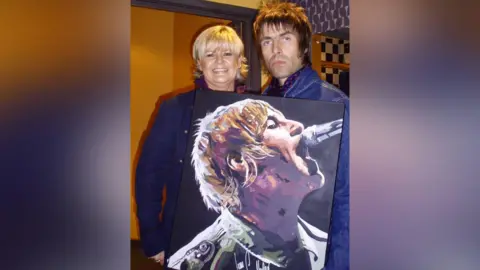 Sue Verity Art A woman with short blonde hair is standing next to a man with black spiky hair and they are holding a portrait of Liam Gallagher from Oasis