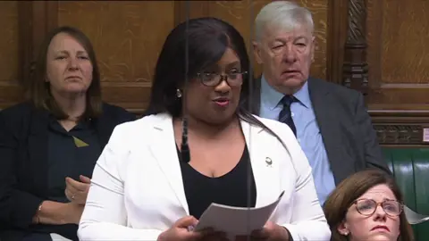 MP Bell Ribeiro-Addy asks a question in the House of Commons. She wears a white jacket on top of a black blouse. Behind her are sat are other Labour MPs, including veteran Labour MP Graham Stringer.