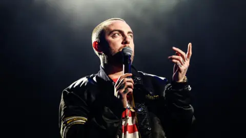 Getty Images Mac Miller di panggung gelap dengan lampu sorot di atas. Dia mengenakan jaket bomber hitam dengan atasan bergaris merah putih dan memegang mikrofon di wajahnya dengan tangan kanannya sambil menunjuk ke langit dengan tangan kirinya. 