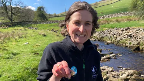 A woman stands close to the camera, wearing a black fleece jacket with a zip front. On the left side of the chest  there is a white logo resembling an  animal emblem. She is holding a crayfish in her hand