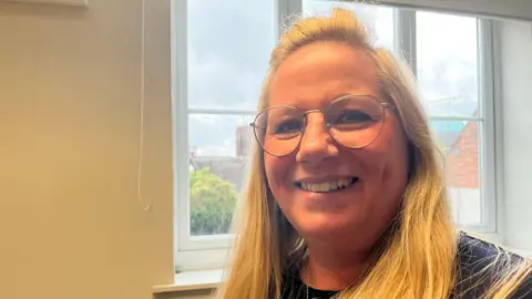 Rebeker Ford has blonde hair and glasses. She is smiling into the camera. Behind her is a window and a cream coloured wall