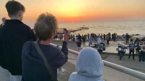 A woman in blue top takes a photo on her phone of the sunrise, looking out to sea, two younger boys either side