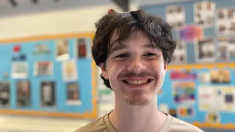 Raven, a 19-year-old man with brown hair and a small beard and moustache smiles at the camera. He is wearing a light brown T-shirt.