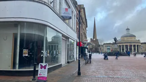 A pedestrianised high street with an empty retail building to the left. There are people in the street and a statue of Robert Blake in the background. There is also a church in the background. The picture was taken on a cloudy day.