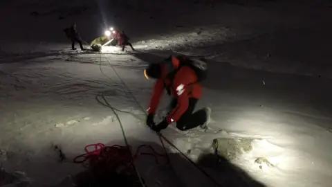 Patterdale Mountain Rescue Team Patterdale Mountain Rescue Team assembling a rope system during a rescue. There is a team member wearing red in the foreground and other team members wearing head torches a few metres away.