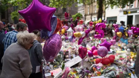 AFP via Getty Images Tributes to the victims of the Manchester arena attacks are shown filling the city centre in 2017, with members of the public looking at a huge number of flowers and pink balloons filling a city centre street.