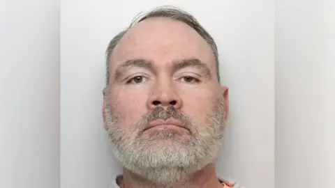 Wayne Stead with short dark hair and grey beard. He is looking at the camera in a police custody photo.