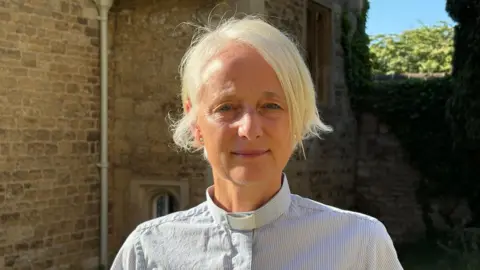 Reverend Clare Hayns, a woman wearing a dog collar, stands outside in a posed photograph. She has short blonde/white hair and she is standing in front of an old stone building in the sun. 