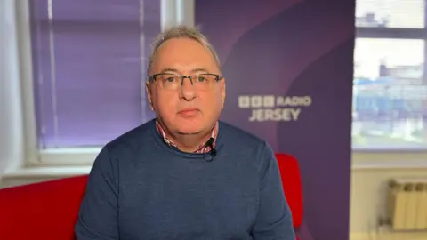 Kevin Keen sitting on the BBC Jersey sofa, wearing a blue jumper and glasses. 