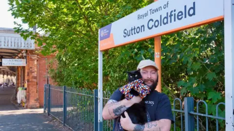 A man is standing on a train station platform holding a black cat. He has a cream cap on, black beard and tattoos. Behind him there is a sign saying "Royal Town of Sutton Coldfield", blue fence and platform. 