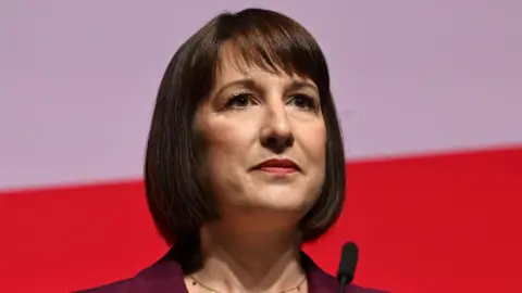 A close up of the chancellor. She has brown bobbed hair. She's wearing a brown jacket and a gold necklace and is standing in front of a mic. The background is white and red.