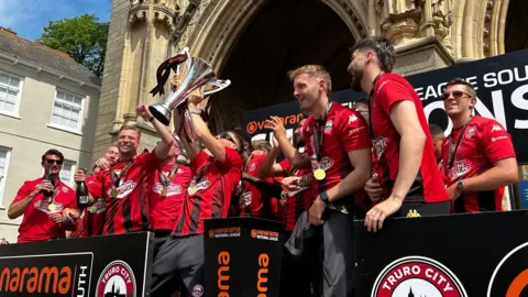 The team lift the trophy while standing on the steps of Truro Cathedral