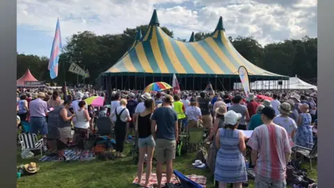 Martin Heath/BBC A large big top with four gables is set against a backdrop of trees. Crowds of people stand beneath it and outside it, wearing summer clothing. There are some colourful flags and umbrellas aroud, and more marquees in the distance.
