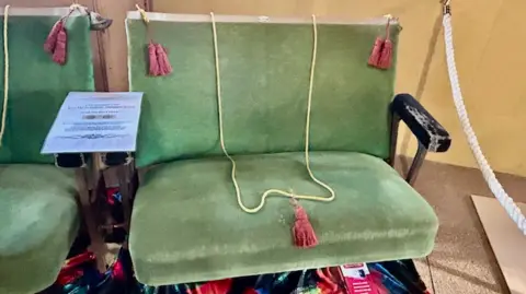 Museum of Bath at Work A close up of two green velvet-covered cinema seats with old wooden handles. There is a thin yellow rope hanging over the chair to prevent visitors sitting on it and pink tassels hanging from the corners of the seat back.