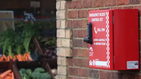 The image shows a bright red Emergency Bleed Control Kit mounted on an exterior brick wall. The kit has instructions printed on the front, including a directive to call 999 and listen carefully to instructions. It features a keypad lock on the left side of the box. In the background, there is an outdoor produce display with vegetables such as carrots and broccoli.