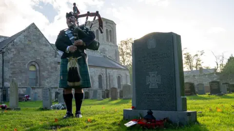 Piper Kevin McLean in full Highland dress, plays his pipes in a churchyard next to the grave of Daniel Laidlaw. The church is in the background and the grave is decorated with poppies.