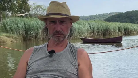 Will is wearing a hat and grey T-shirt as he sits on the ferry with the river behind him.