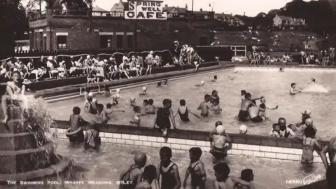 Black and white photo of several people, young and old swimming in the Lido. Picture is undated. A cafe stands near to the pool.
