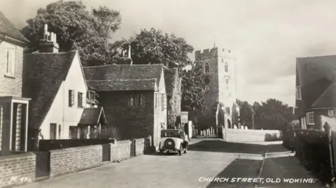 Postcard Traders Association A black and white photograph of Church Street in Old Woking, showing a car parked outside a house and the church in the background.