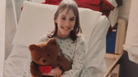 Jessie White Jessie White when she was 11-year-old. She sits on a hospital bed with a hospital gown on. She is holding a brown teddy bear and smiling at the camera. 
