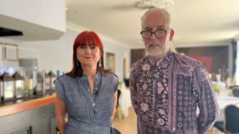 Steve Cuddy and Gillian Etherdige stand together in the dining room of their hotel. To their left is a bar and behind them are tables and chairs with white tablecloths on top. Etheridge has bright red hair and wears a denim dress. Cuddy has grey hair and beard, is wearing glasses and a purple and navy shirt.