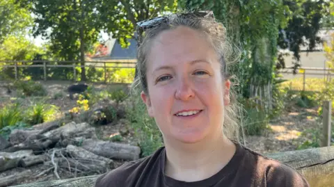 A person wearing a Jersey Zoo and Durrell T-shirt is sitting beside a wooden fence in a leafy outdoor enclosure. Trees, logs and plants can be seen in the background, with a dark animal visible in the distance.