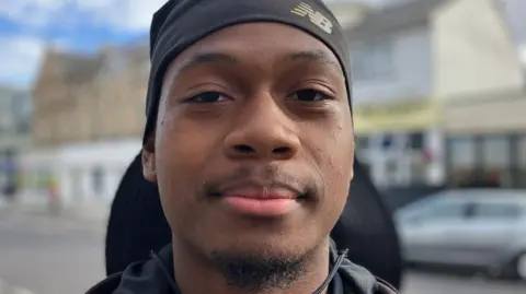 Kaleb, 19, from Tottenham is wearing a black hat which covers the back of his head. He is in a street which has been blurred to focus on his face.