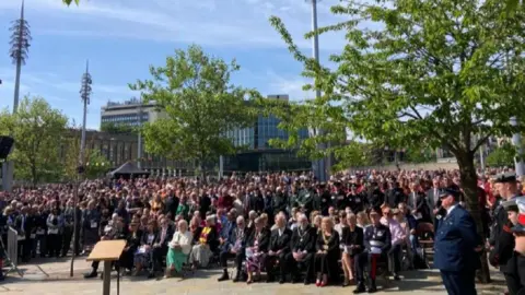 BBC/Cathy Killick Hundreds of people are sat or stood in rows a memorial setting outside.