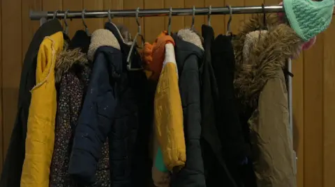 A clothes rack is full of coats with some yellow and blue ones standing out. There is a green hat hanging from the end of the rack and a brown coat with a furry hood next to it.