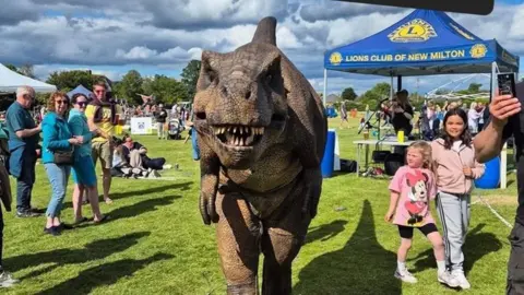 Large green and grey model of a Tyrannosaurus Rex looking straight at the camera with its mouth open and teeth visible. He is standing on grass alongside adults and children at an event. A blue gazebo with yellow writing "Lions Club of New Milton" is visible to the right. Another light-coloured gazebo can be seen to the left.  A young child in a pink T-shirt with a cartoon mouse on the front is looking at the dinosaur.