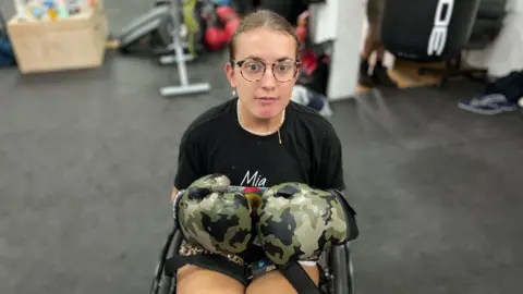 Mia in her chair and wearing camouflage boxing gloves 