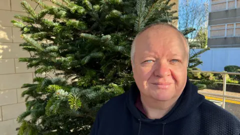 John Devine/BBC Richard, a man, wearing a black hooded jacket, standing in front of a green tree, smiling for camera.
