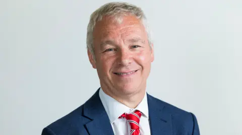 Reading Borough Council Councillor John Ennis smiles at the camera in his official council photo. He has light grey hair and wears a blue jacket, white shirt and red tie.