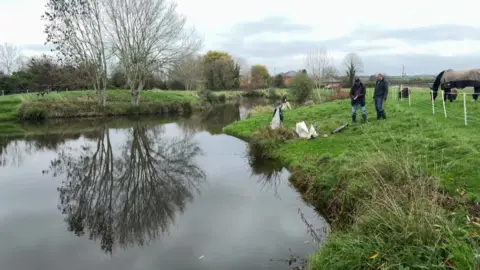 Chris McHugh/BBC A wide shot of a small man-made fishing lake, surrounded on all sides by grassy banks. Two men can be seen standing near the right-hand bank. Trees are in the background