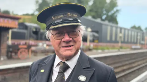 A white-haired man wearing a railway cap that says "station master", a black blazer, black tie and white shirt, standing in front of a railway yard and track.