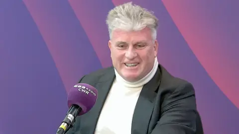 A man in a grey jacket and white polo neck jumper in a radio studio. He is talking into a radio microphone with a purple head and BBC Radio CWR written on it. Behind him is a purple and pink wall.