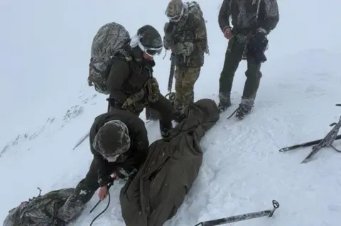 Royal Navy A group of marines dressed in green clothing during exercise on Ben Nevis. They are standing in snow and ice axes are laid on the ground.