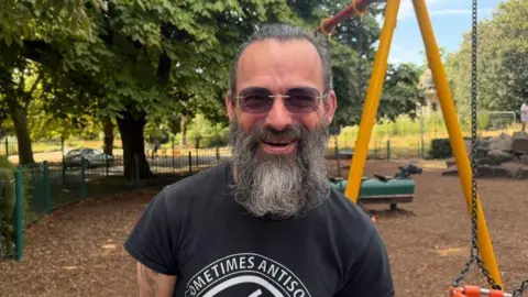 Jasmine Lowe/BBC News A man in a children's park on a sunny day. He has a black T-shirt on and he has very long hair. He also has a long beard and it's black with grey hairs in. He's wearing sunglasses and looks smiley.