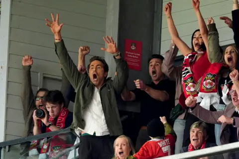 PA Media Ryan Reynolds and Rob Mac can be seen in the stands at Wrexham AFC passionately react in celebration to something on the football pitch that cannot be seen. They appear to be shouting and raising their hands. They are surrounded by other fans making similar reactions. 