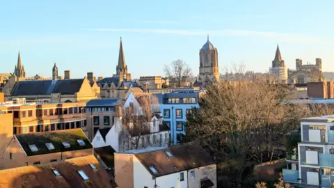 Lucie Johnson A city skyline under a clear blue sky on a winter's day. There are streets of homes and flats in the foreground with bare trees. In the distance you can see the spires of the Oxford colleges.