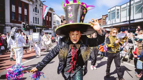 Andrew Billington A man in an oversized top hat is looking at the camera, as a marching band is seen playing behind him. He is in the middle of a street in Longton, Stoke-on-Trent.
