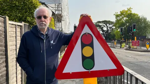 An elderly man in sunglasses wearing a dark blue hooded jumper. He is stood beside a road looking into the camera. He has his left hand on top of a triangle road sign showing a set of traffic lights
