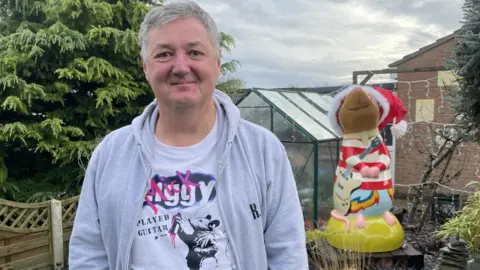 Jon Seth - a man wearing a grey hoodie and rat t-shirt honouring Mick Ronson - standing in front of a giant red and white striped rat sculpture holding a guitar. The sculpture has a red Santa hat on its head.