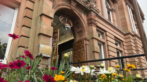 An image from a flower bed looking up at the entrance of the Ewart and its bronze ornate gates